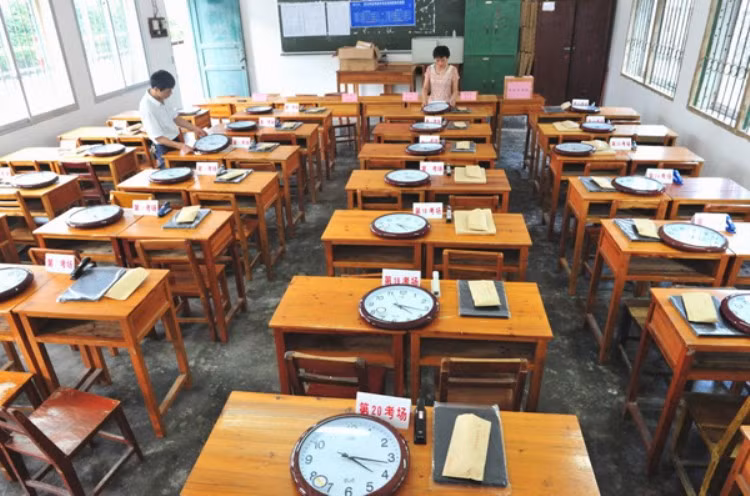 Cận cảnh phòng thi đang được "set up". Mỗi một bàn sẽ có một chiếc đồng hồ đã được căn giờ giống nhau, đề thi và giấy nháp. (Ảnh: Kenh14)