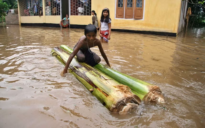 Một cậu bé ngồi trên một chiếc bè chuối bên ngoài ngôi nhà bị ngập nước ở vùng ngoại ô thành phố Guwahati.