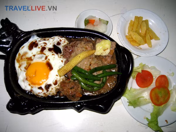 35. Bò bít tết: Thịt bò xắt miếng to và dày, sau đó đem áp chảo cho chín tái rồi tưới thêm gia vị. Món này dùng với tương ớt, xì dầu hoặc nước sốt đặc biệt, với vài miếng xà lách, cà chua, chưa leo...Ăn đâu: Bò Bít Tết Hoả Diệm Sơn – 331 Nguyễn Trãi, Q.1, TP.HCM ...