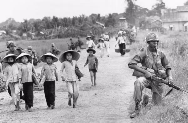 Sư đoàn lính Mỹ kiểm soát phụ nữ và trẻ em Việt Nam đi học từ Bến Cát trở về nhà ở làng Xuân Điền. Bức ảnh được chụp vào tháng 12 năm 1965.